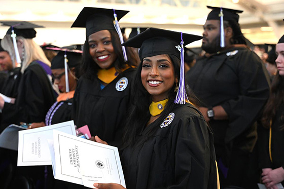 Two graduating students in robes hold up their degree and smile for a photo