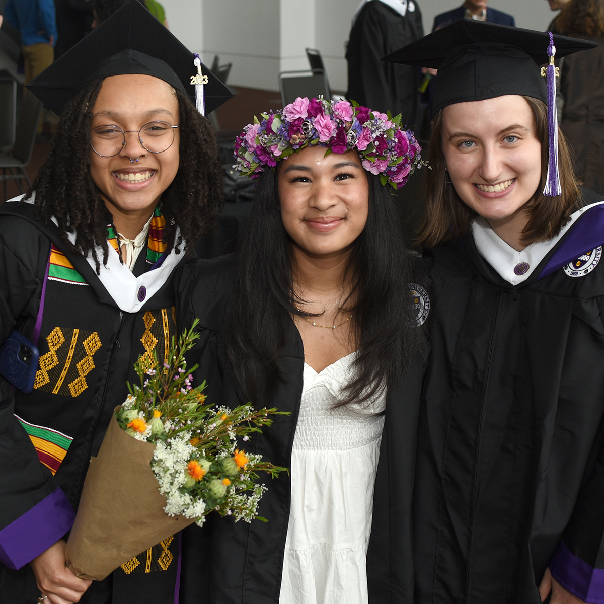 Three graduating students in robes, two in traditional caps and one wearing a beautiful flower crown, pose for a photo