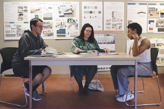 A professor and two students are seated at a table, discussing projects