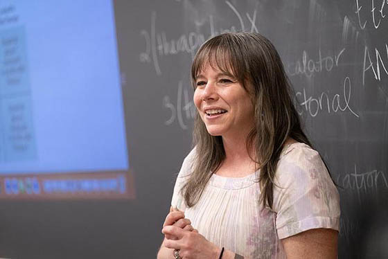 Photo of a professor smiling at the front of a classroom, instructing students off-screen with a chalkboard behind her