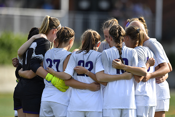 Photo of Chatham University's women's soccer team in white and purple uniforms huddles on the field during a match. 