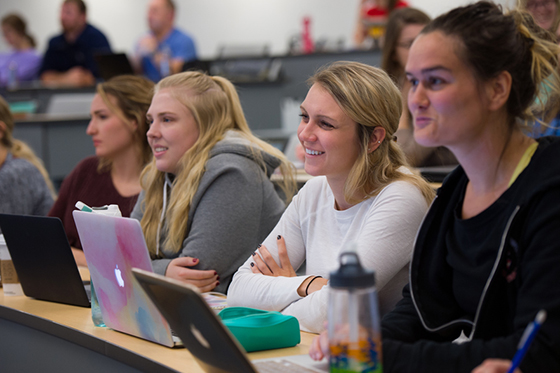Photo of four female Chatham University students smiling in a lecture hall, with their laptops open in front of them