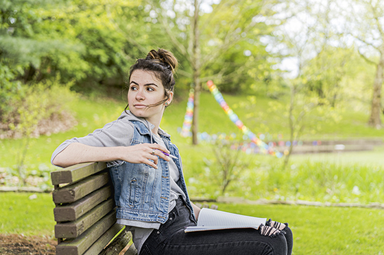 Photo of a Chatham University student sitting on a bench outside writing in notebook on lap. 