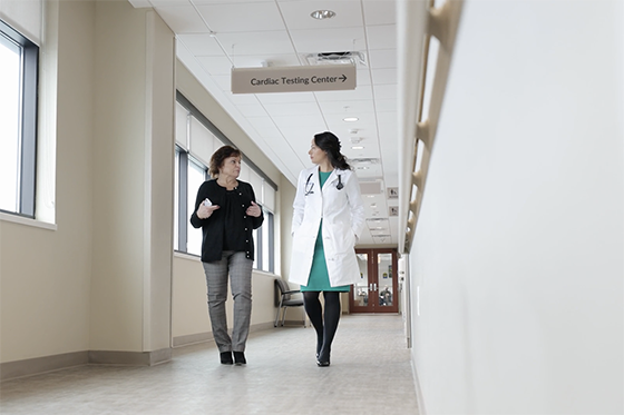 Photo of a doctor in a teal dress and white lab coat speaking with an older female patient as they walk down a hallway together