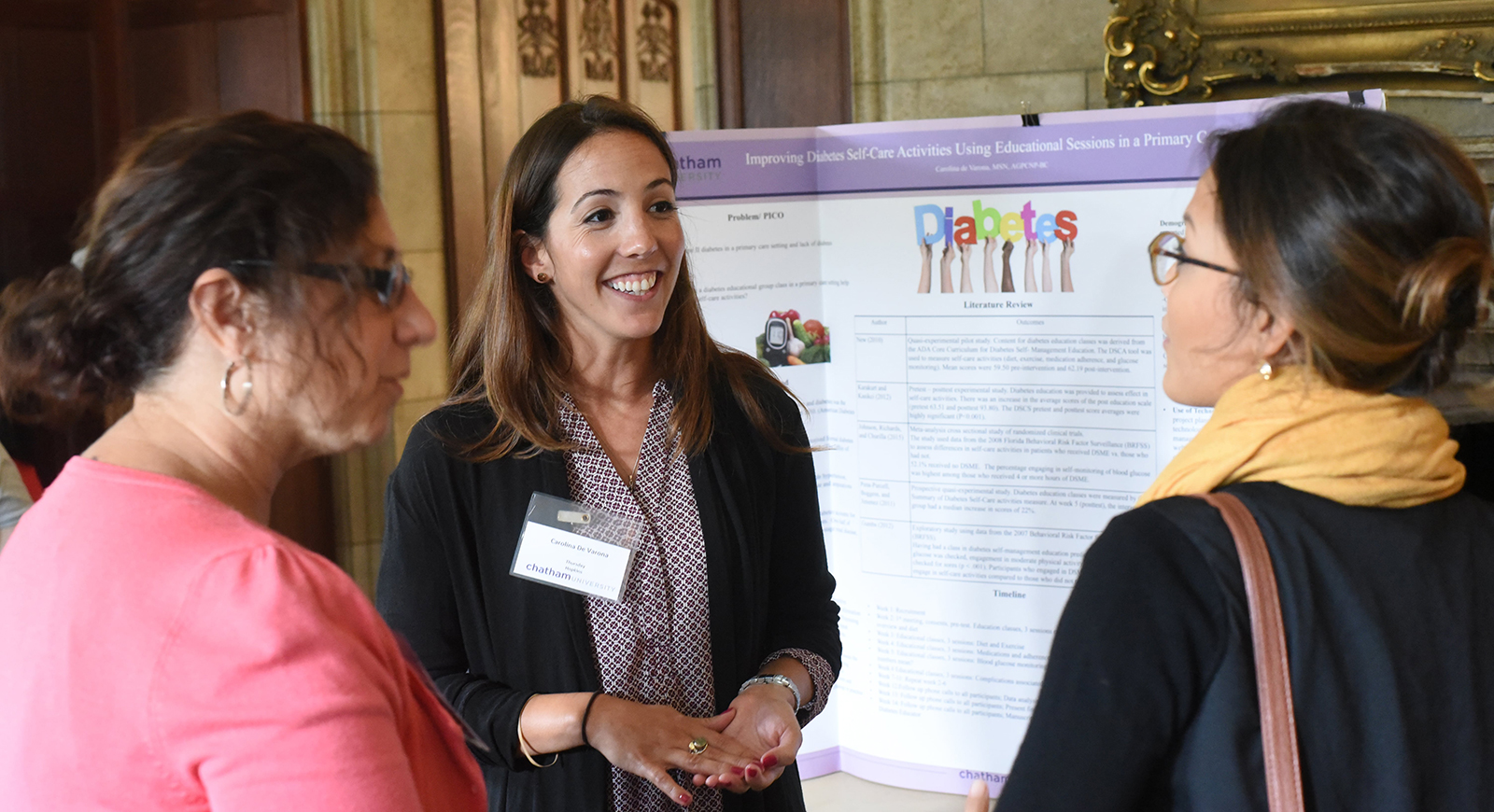 Carolina De Varona speaks to two women wearing business casual attire in front of a poster presentation. 