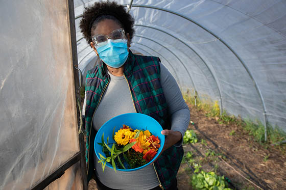 Photo of a masked Chatham University student holding a blue bowl of harvested produce, at the entrance to a greenhouse on Eden Hall Campus