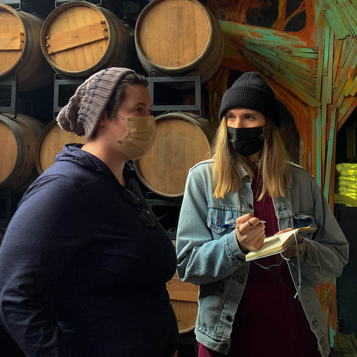 Photo of two people wearing masks, one with a notebook and pen in hand. Behind them there are barrels.