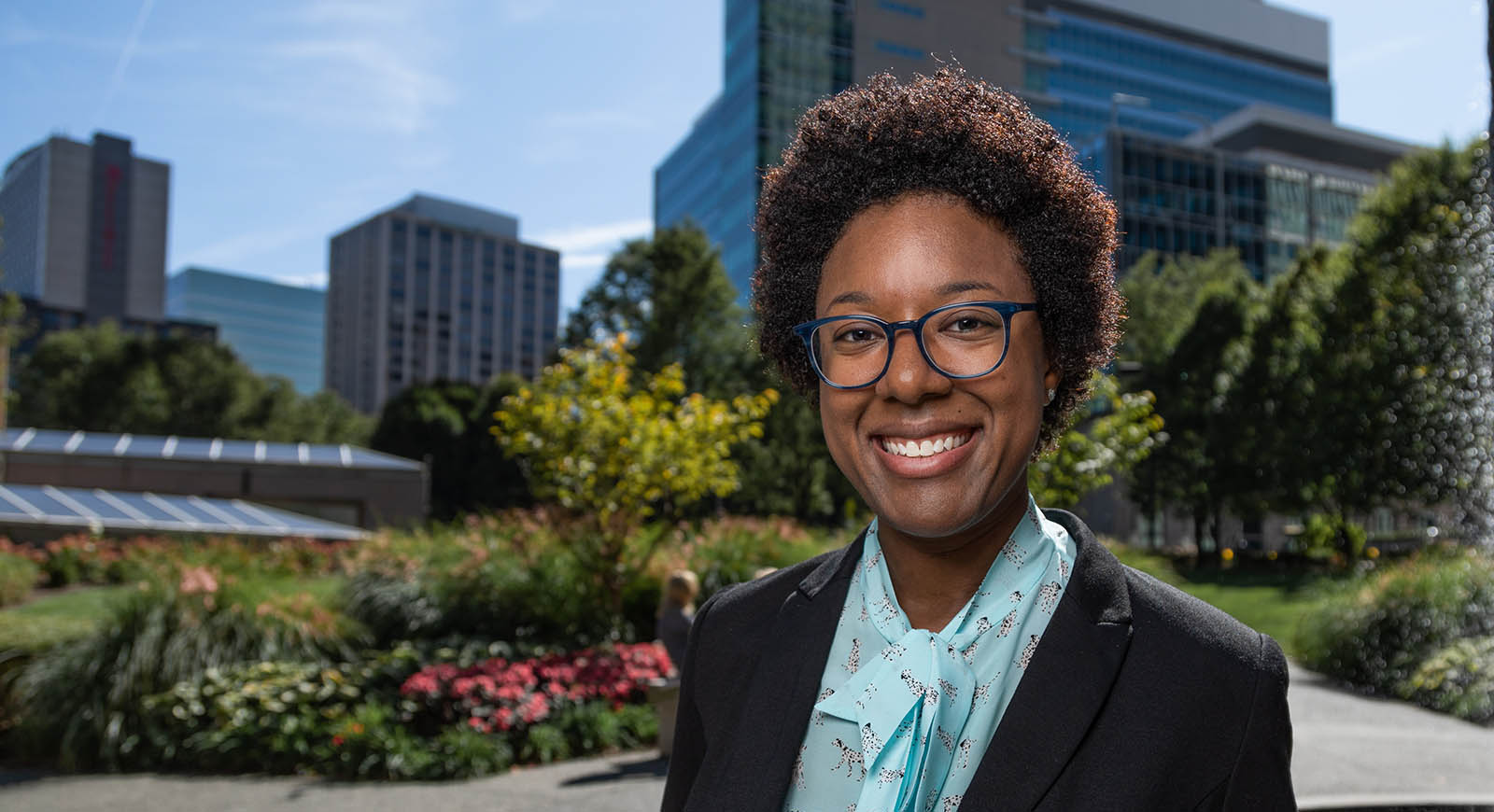 Photo of a smiling woman in professional clothes, posing in front of buildings and greenery in downtown Pittsburgh