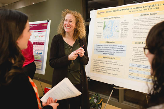 Photo of a smiling woman in front of a poster presentation, speaking with two women facing her