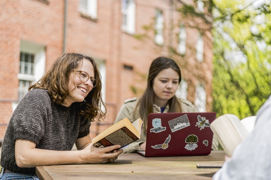 Photo of three Chatham University students sitting at an outdoor picnic table reading and working on a laptop. 