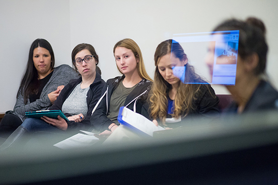 Photo of five Chatham University psychology students sitting behind glass in an exam room. There is a reflection of a screen on the glass. 