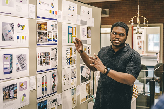 Photo of a Chatham University design student presenting in front of a wall of printed design material. 