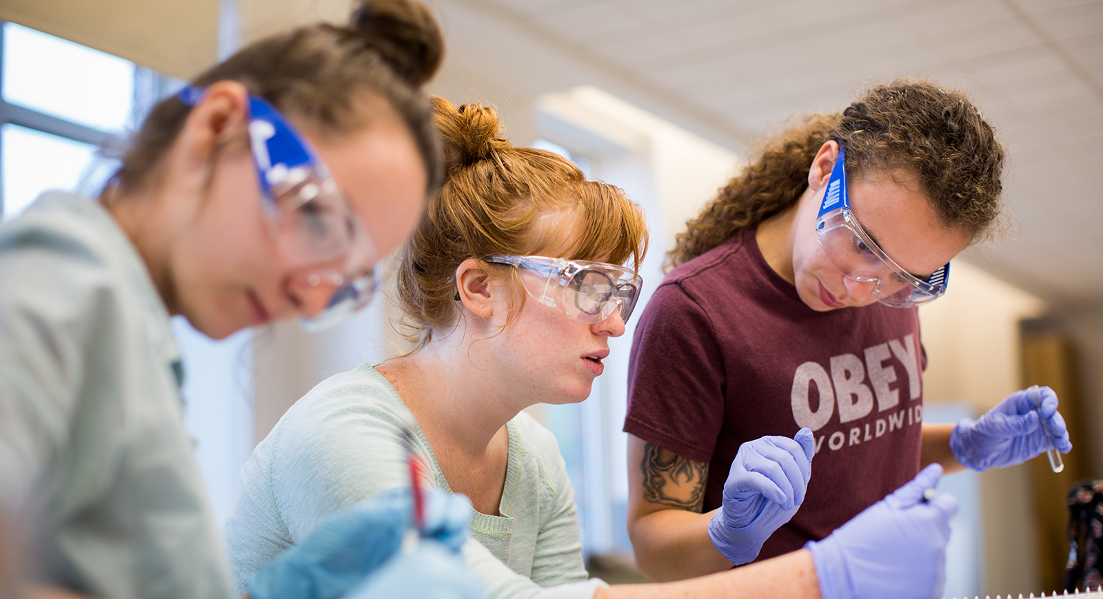 Photo of three female Chatham University biochemistry students wearing blue gloves and goggles, working in a lab holding glass test tubes. 