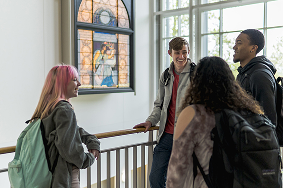 Photo of a group of four Chatham University students laughing after class in brightly lit academic building with a stained glass window. 