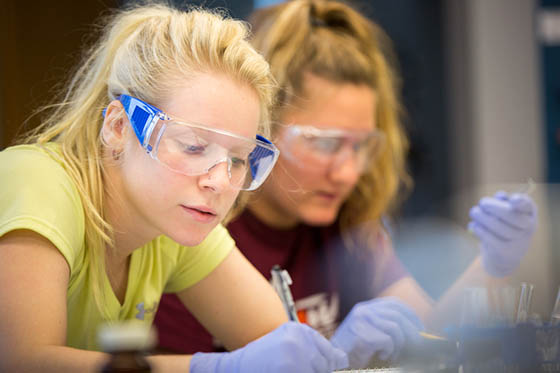 Photo of two female Chatham University students wearing protective goggles and writing at a lab table