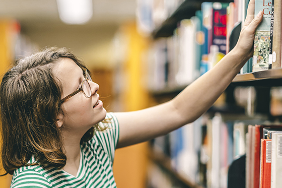 Photo of a female Chatham University student in green and white striped t-shirt reaching for a book in the stacks at Jennie Mellon King library. 