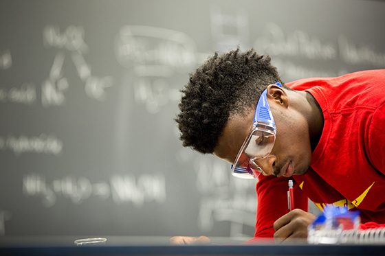 Photo of a Chatham University student wearing a red shirt and protective goggles in a lab, writing with a pencil. 