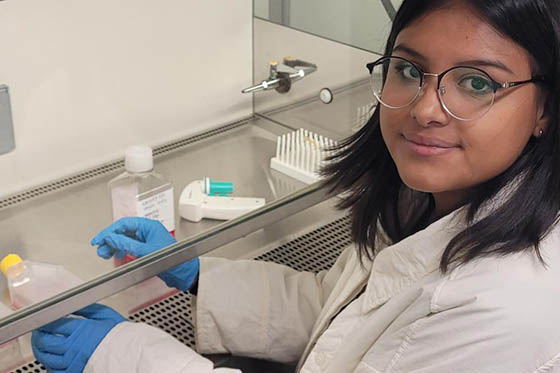 Photo of a young woman with glasses, wearing a lab coat and blue latex gloves, poses for a photo while working in a lab