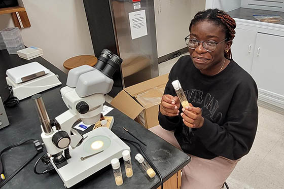 Photo of a young Black woman wearing khakis and a black sweatshirt, sitting on a stool in front of a microscope and holding a test tube