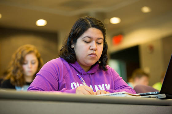 Photo of a Chatham University student writing in a notebook, in a lecture hall. Other students are sitting around her.