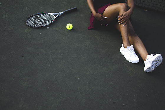 Photo from above of a woman adjusting her leg, sitting next to a tennis racket and ball. 
