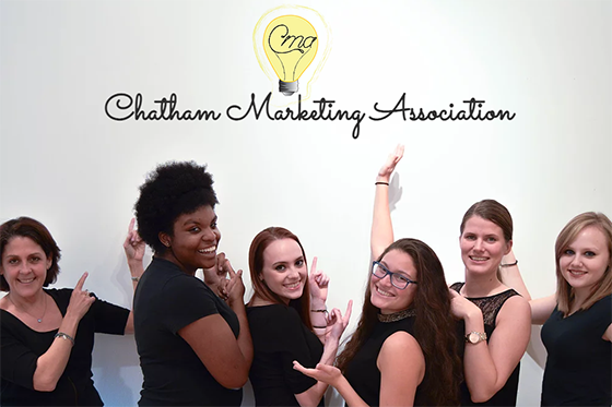 Photo of a group of students and professor, holding their hands up in Vana White style to showcase the Chatham Marketing Association logo