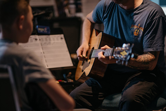 Photo of a man with tattoos teaching acoustic guitar to a child sitting across from him