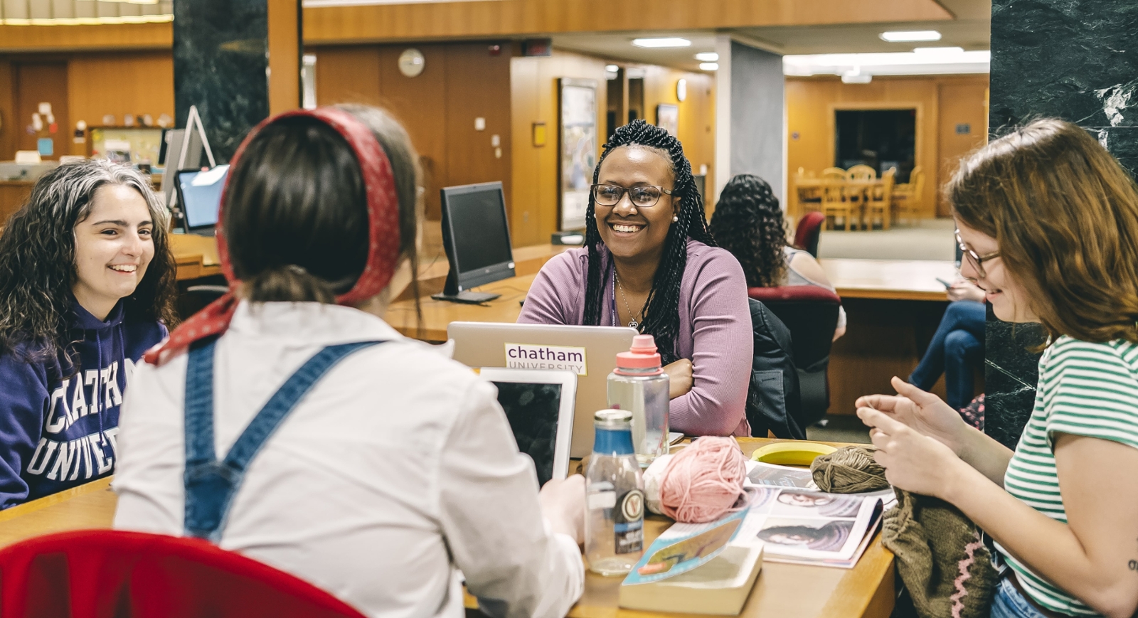 Photo of four female Chatham University students sitting at a table in Jennie Mellon King Library talking with laptops, notebooks, and knitting. 