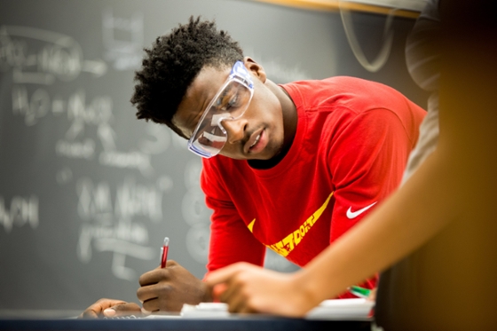 Photo of a student in protective lab gear writing down experiment observations in his notebook
