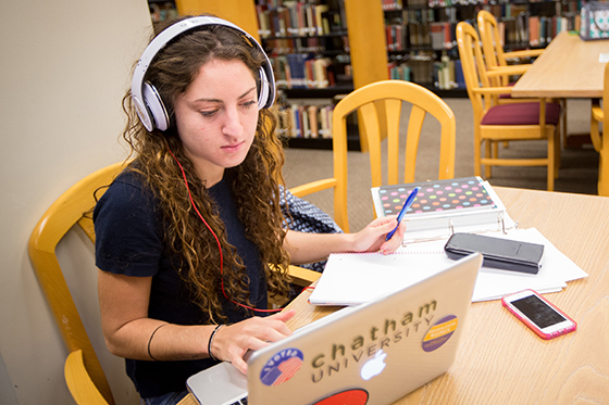 Photo of a Chatham University student sitting at a table in Jennie Mellon King Library studying on a laptop and writing in a notebook with a calculator. 