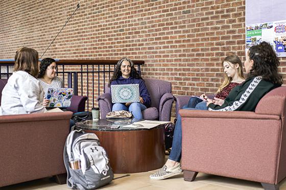 Photo of five Chatham University students sit together in chairs outside of classroom working on laptops and talking