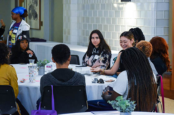 Photo of four students sitting around a table in the library, talking and laughing, with laptops and books and knitting on the table