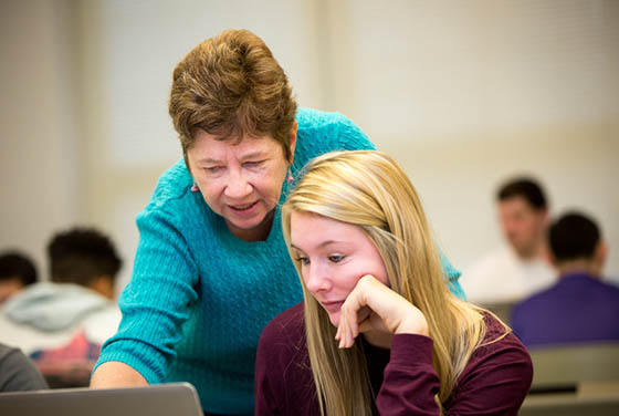 Photo of an older female faculty member leaning over a Chatham University student and pointing to a computer