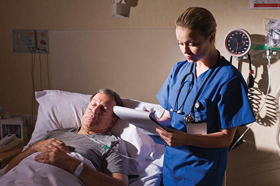 Photo of a nurse in scrubs, writing on a clipboard next to a sleeping patient