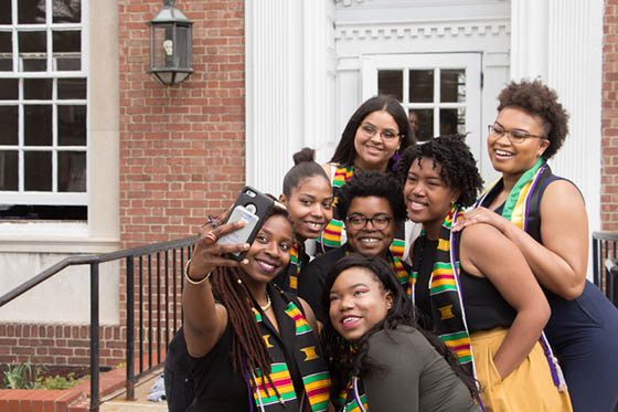 Photo of seven BIPOC Chatham University students in gramulticultural graduation stoles, posing for a selfie in front of James Laughlin Music Hall on Shadyside Campus