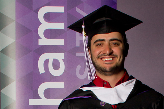 Photo of a smiling male student in graduation cap and gown, posing in front of a sign reading Chatham University