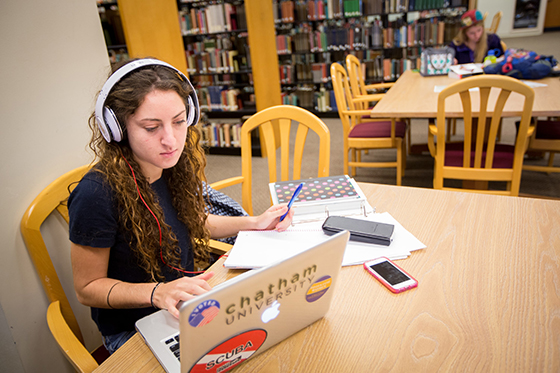 Chatham University student sits at Jeanie King Mellon Library, studying at a table while wearing headphones and working on her computer. 