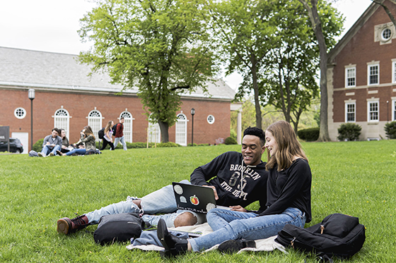 Two Chatham University students sit on a green grass academic quad, working together on a laptop. 