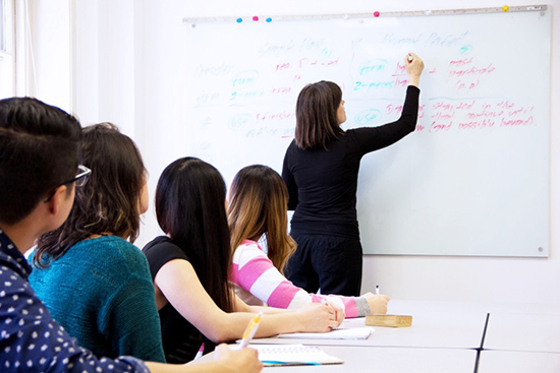 Photo of students watching a professor write notes on a whiteboard. 