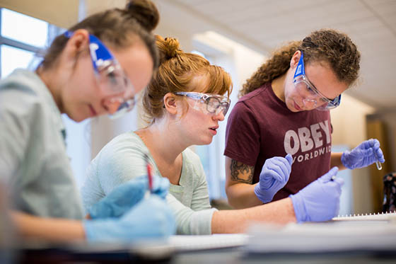 Photo of three female Chatham University students in protective goggles and gloves working in a lab