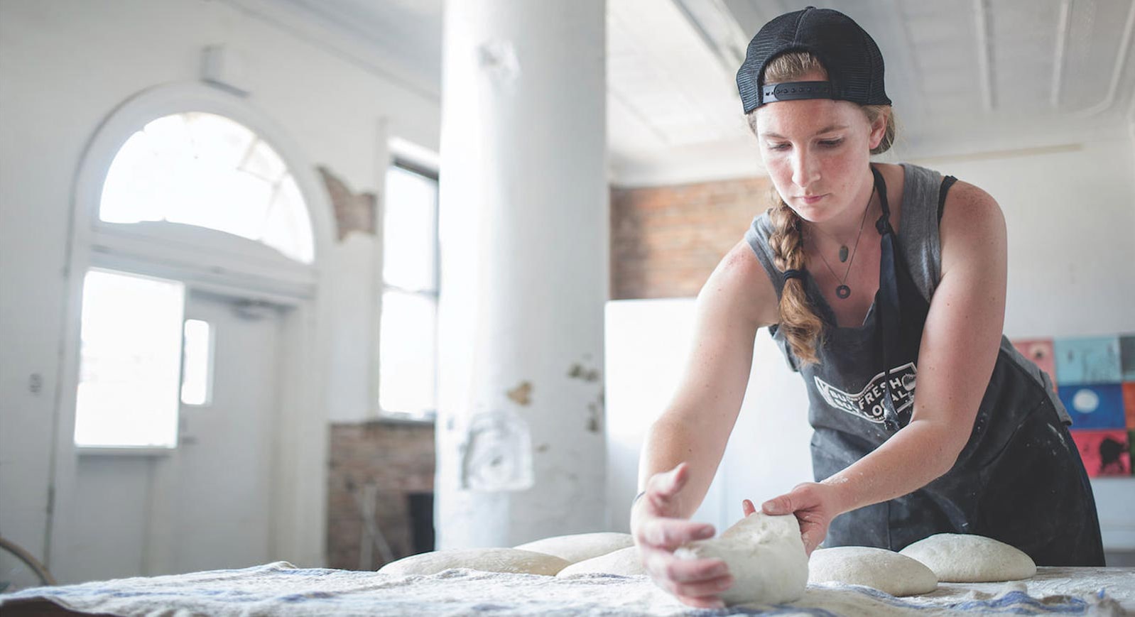 Photo of a woman arranging dough on a floured table in a light filled open room while wearing a backward baseball cap and apron. 