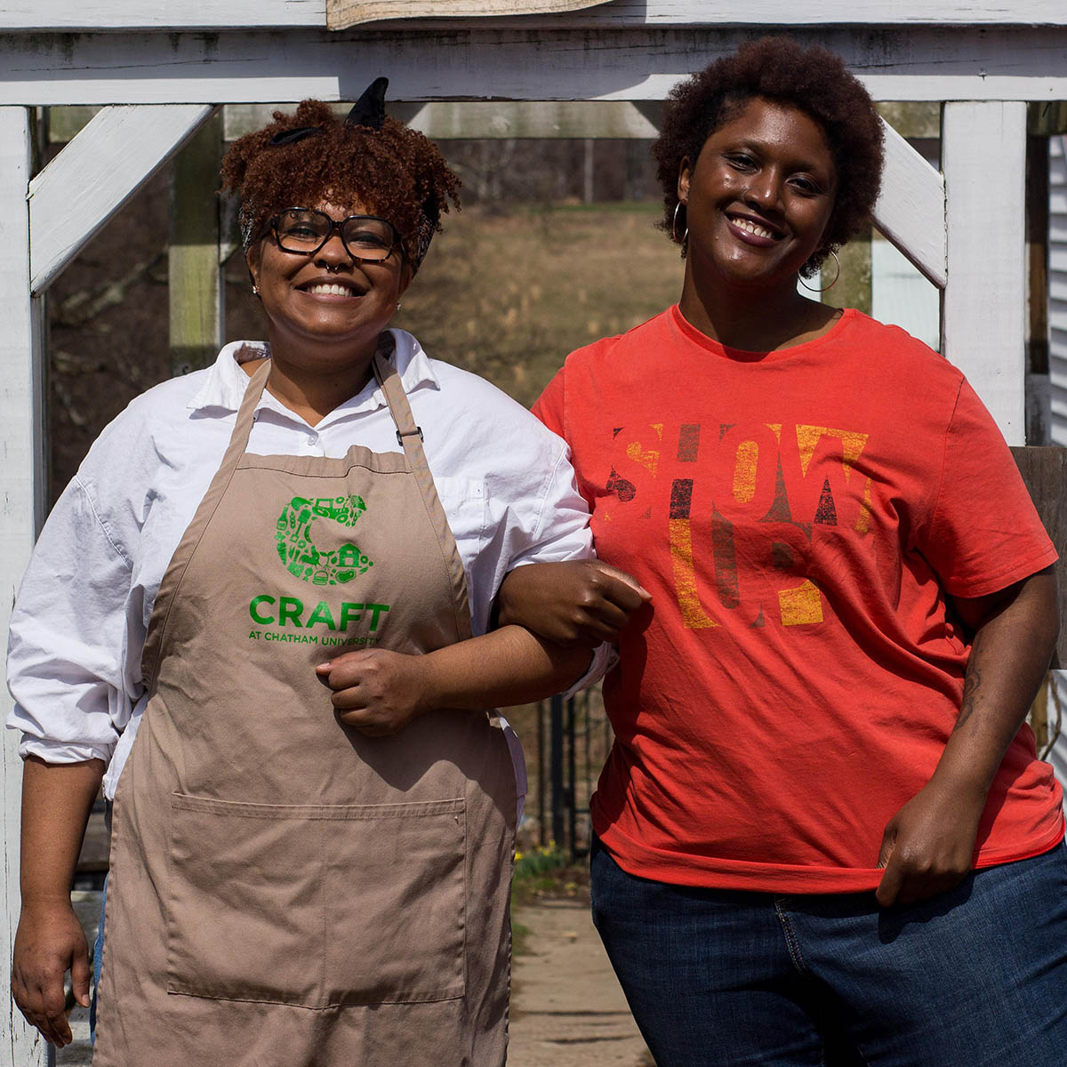 Photo of two smiling Black women linking arms. One is wearing a CRAFT apron.