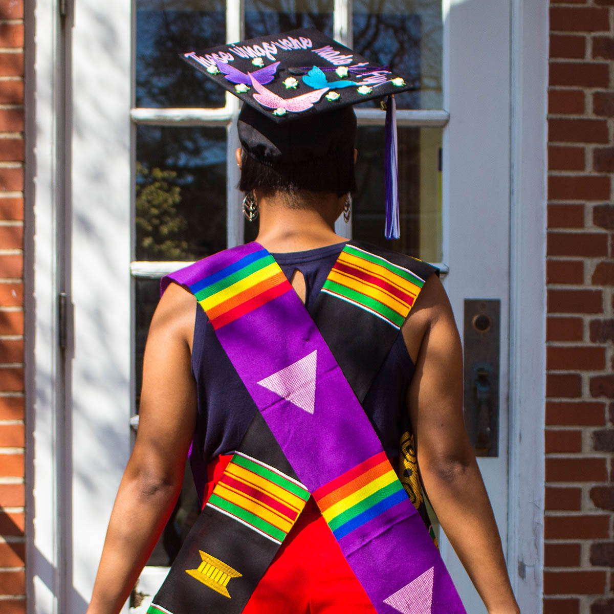 Photo of a student facing away from the camera, wearing a rainbow graduation stole and a graduation cap reading "these wings were made to fly"