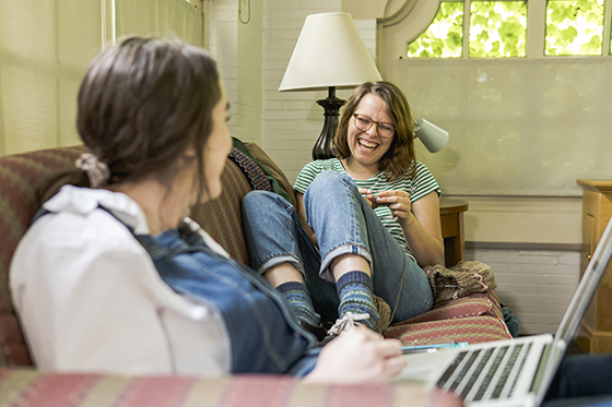 Two Chatham University students sit on a couch laughing and smiling while doing homework together.
