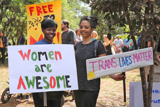 Photo of two young women holding signs reading Women Are Awesome and Trans Lives Matter