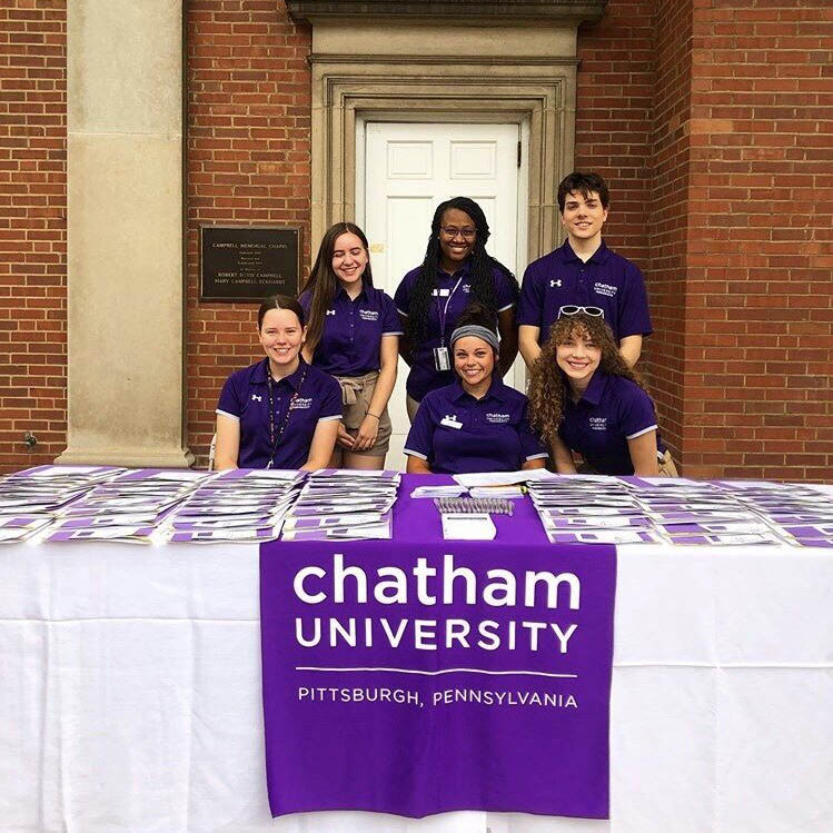 Photo of six Chatham University students in purple shirts, working at a table outside on Shadyside Campus