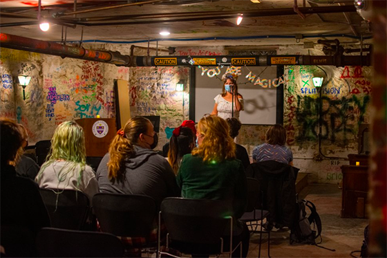 Photo of an event in Rea Coffeehouse, with students seated facing a stage with a person speaking into a microphone