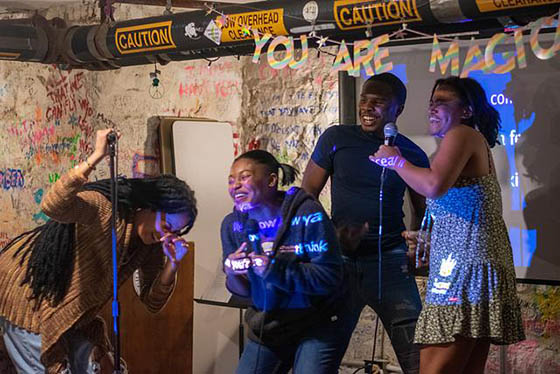 Photo of four Black students laughing and sining on stage with microphones during BIPOC karaoke night. There is a sign behind them that says YOU ARE MAGIC.
