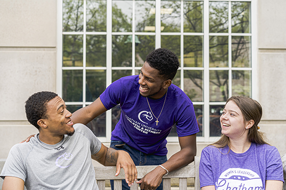 Photo of three Chatham University students sitting outside on a bench laughing and smiling together. 