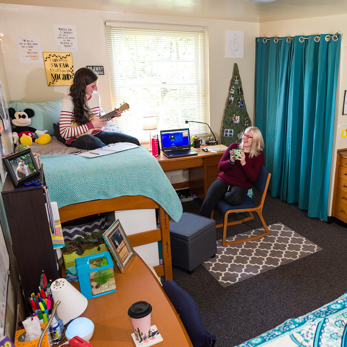 Photo of two roommates laughing and hanging out in their dorm room. One of them is playing a ukulele.